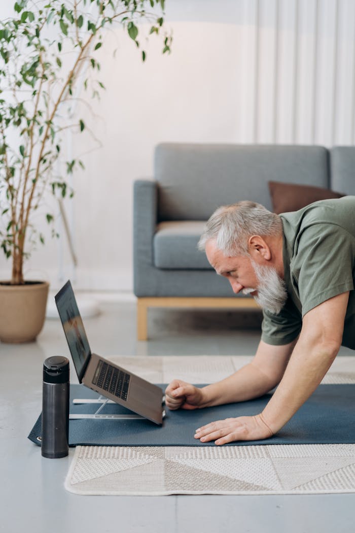 Offerings Elderly man engaging in yoga exercise at home using a laptop for guidance.